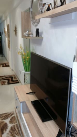 A modern living room with a flat-screen TV on a wooden stand, a white cabinet below, and a decorative plant to the left. - Beach Front Luxury Living Apartment (Port El Kantaoui)