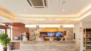 A hotel reception area with a wooden reception desk, two receptionists, a digital screen, and a potted plant. - Greentree Inn Qufu High Speed Railway Station (Jining)