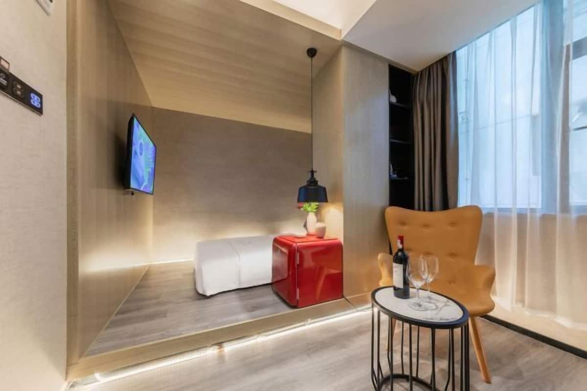 Ein modernes Hotelzimmer mit Flachbildfernseher an der Wand, einem roten Hocker, einem runden Tisch mit einem Weinglas und einer Weinflasche sowie einem großen Fenster mit leichtem Vorhang.