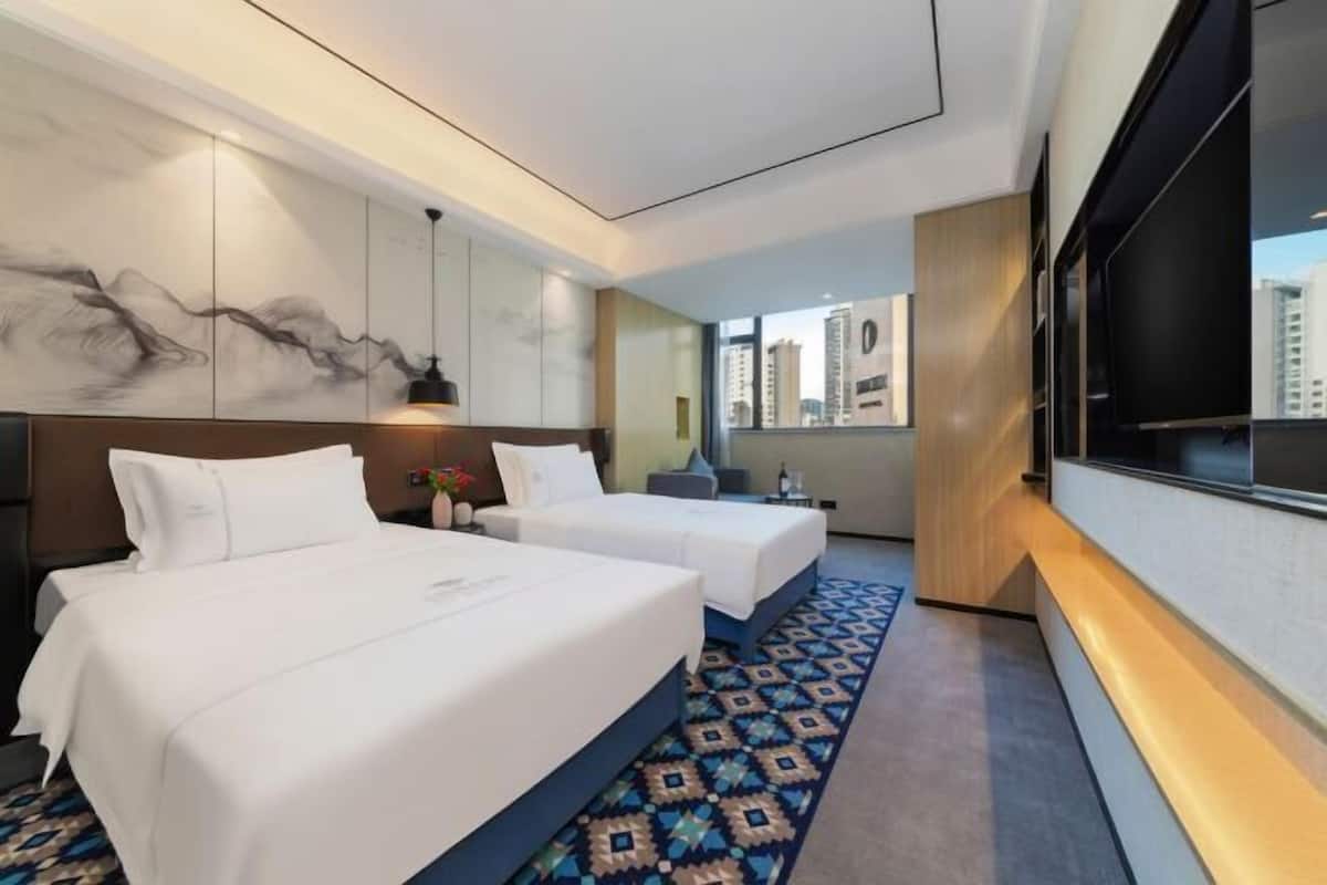 Ein Hotelzimmer mit zwei Betten, einem großen Fenster mit Blick auf die Stadt und einem wandmontierten Fernseher.