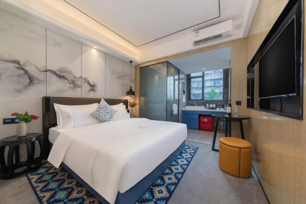 Ein modernes Hotelzimmer mit einem großen Bett, einem kleinen Schreibtisch, einem Fernseher und einem durch eine Glaswand abgetrennten Badezimmer.