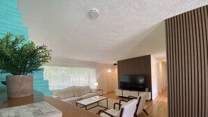 A modern living room with a sofa, coffee table, and chairs. There is a TV on a wooden stand, a potted plant, and a staircase with blue steps.