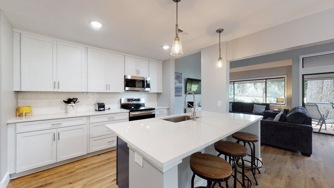 A modern kitchen with white cabinetry, a large island with stools, a microwave, and a sink.