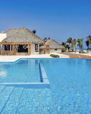 A clear blue pool with wooden deck areas and thatched-roof structures. - Apartamento Trendy, Airport Transfer (Punta Cana)