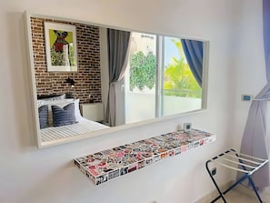 A bedroom with a bed, a large mirror, a window with curtains, a wall-mounted shelf with stickers, and a drying rack. - Apartamento Trendy, Airport Transfer (Punta Cana)