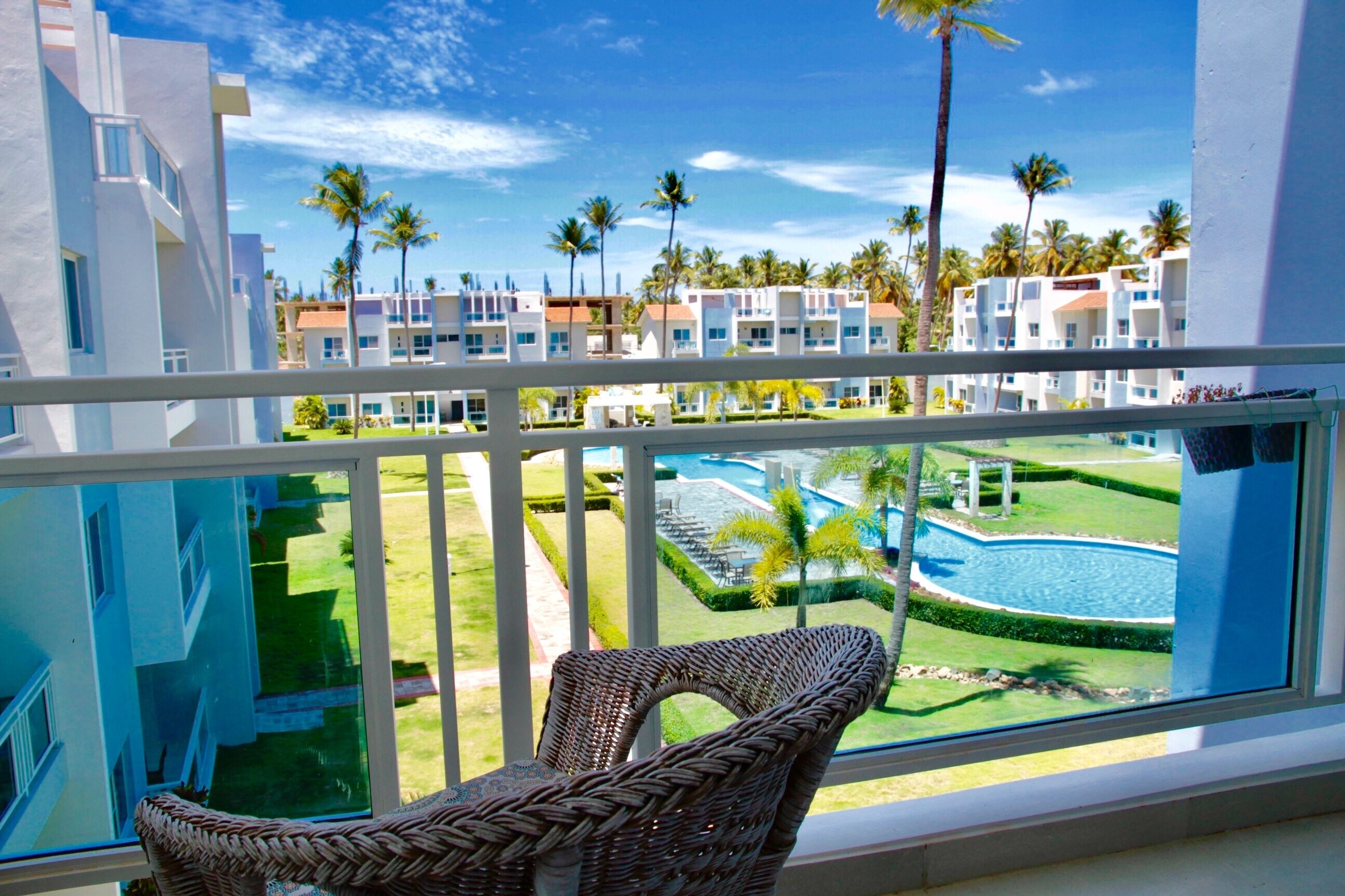 A balcony with a wicker chair, overlooking a pool area with palm trees and a building complex.