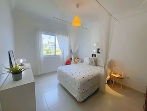 A bedroom with a bed, a bedside table, a potted plant, a window with curtains, and a hanging light fixture.