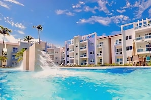 A swimming pool with a water feature, surrounded by multi-story buildings with balconies and outdoor seating.