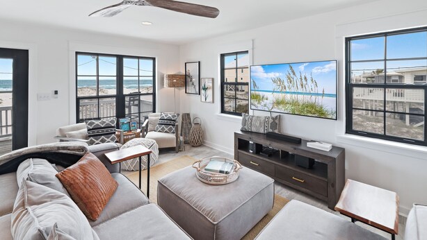 Un salón amplio con un gran ventanal con vista a la playa, un ventilador de techo y un televisor de pantalla plana.