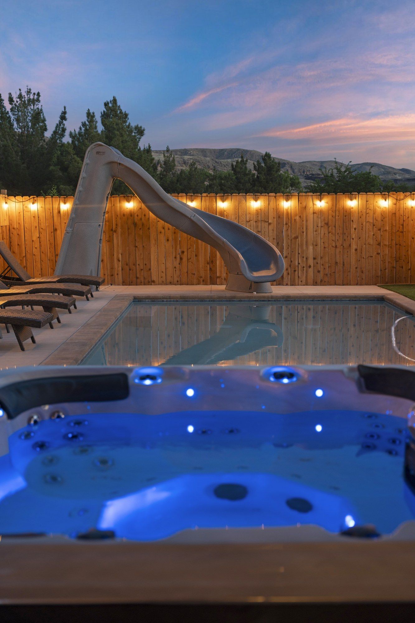 A hot tub with a wooden deck and lounge chairs, surrounded by a wooden fence with lights.
