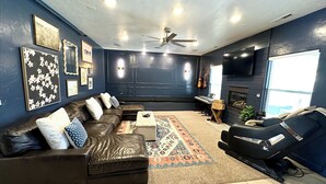 A modern living room with a dark blue wall, a leather sofa, a coffee table, a gaming chair, a piano, and a fireplace.