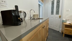 Une cuisine équipée d’une machine à café, d’une bouilloire et d’un évier. On y trouve un meuble en bois et une porte ornée de panneaux de verre décoratifs.