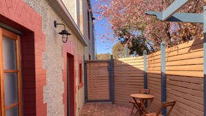 Une étroite allée bordée de briques, avec une porte en bois, une petite table en bois et deux chaises, et une clôture en bois.