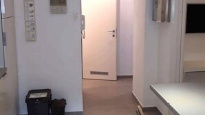 Un pasillo estrecho con una puerta blanca al final, un bote de basura y una pequeña mesa con una silla.