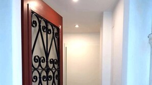 Un pasillo con una puerta de madera que presenta una reja metálica decorativa. La puerta es blanca con un marco rojo y hay un accesorio de iluminación encima. El pasillo conduce a otra puerta blanca al final.