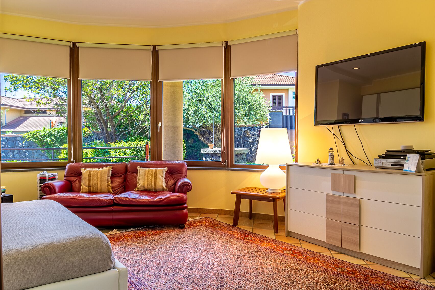 Ett hotellrum med en röd soffa i läder, ett litet träbord med en lampa, en platt-TV som är monterad på väggen och ett stort fönster med utsikt över grönska och byggnader utanför.
