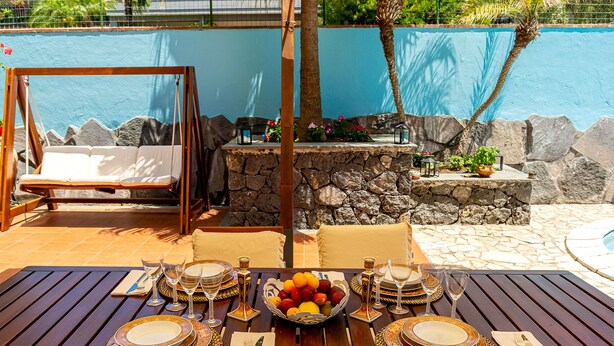 Un comedor con una mesa dispuesta para cuatro personas, rodeado por un muro de piedra y una piscina.