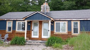 Una casa pequeña de un solo piso con fachada azul oscuro, una chimenea de piedra y un porche con dos sillas Adirondack.