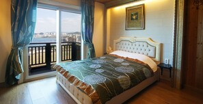 A bedroom with a bed, a view through a balcony door, a framed picture on the wall, and a nightstand.