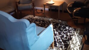 Een woonkamer met een blauwe bank, een grijze fauteuil, een kleine tafel met een lamp, en twee ingelijste foto's aan de muur.