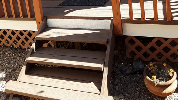 Een houten terras met treden naar een deur, een kleine bloembak met bloemen en een houten schutting.