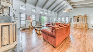 A spacious living room with hardwood floors, a red sofa, a wooden cabinet, and a ceiling fan. - Spacious Ruidoso Retreat: Game Room & Pool Access! (Ruidoso)