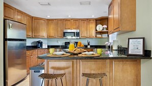 A kitchen with wooden cabinets, a stainless steel refrigerator, a microwave, and a coffee maker. There are two woven stools at the island and a framed welcome sign on the wall. - **SET SAIL @ COCONUT MALLORY** Condo + Last Key White Glove Service... (Key West)