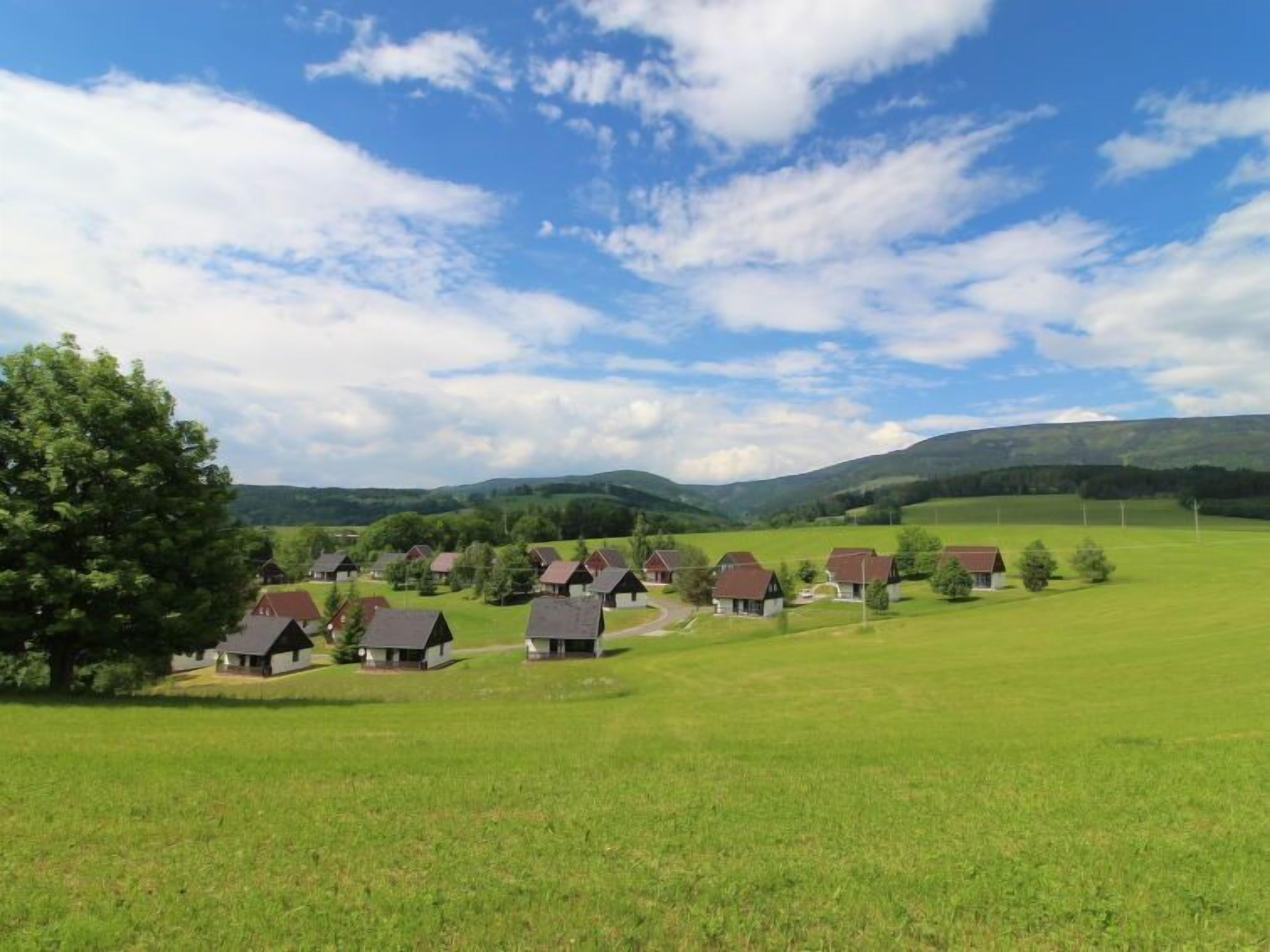 Een landelijk dorp met traditionele huizen, een groot groen veld en glooiende heuvels op de achtergrond.