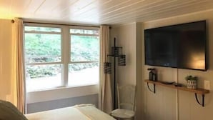 Ett rum med en säng, en tv som är monterad på väggen, ett fönster med gardiner och en hylla med en växt.