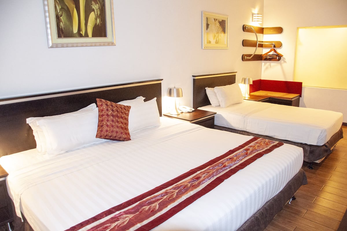 Habitación de hotel con dos camas, cada una con ropa blanca y un corredor decorativo rojo. Hay lámparas de pared y un cuadro sobre las camas.