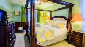 Uma cama de dossel com colcha estampada, um aparador com gavetas, uma mesa de cabeceira com abajur e um ventilador de teto.