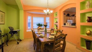 Uma sala de jantar com mesa de madeira posta para seis pessoas, rodeada por cadeiras, um candelabro e vista para a cozinha através de um arco.