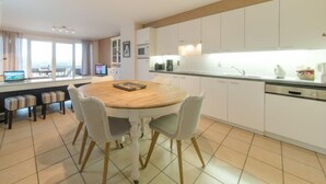 A modern kitchen with white cabinets, a wooden dining table, and a view of the city through large windows. - D’Heyezicht with parking by Interhome (Bredene)