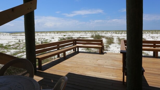 Une terrasse en bois, dotée de bancs, surplombe une plage de sable où la végétation est clairsemée.