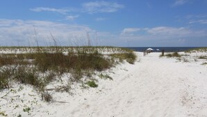 Un sentier de sable qui longe la plage et mène à une structure en bois surmontée d’un parasol bleu.