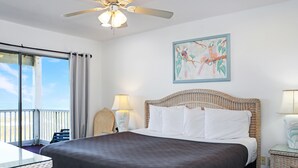 A hotel room with a large bed, a ceiling fan, a painting on the wall, and a view of the ocean through a sliding door. - Beachfront, Quiet shoreline ~ Flamingo Dunes (St. Joe Beach)