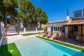 A swimming pool with lounge chairs, a pergola with a bench, and a stone building with a tiled roof.