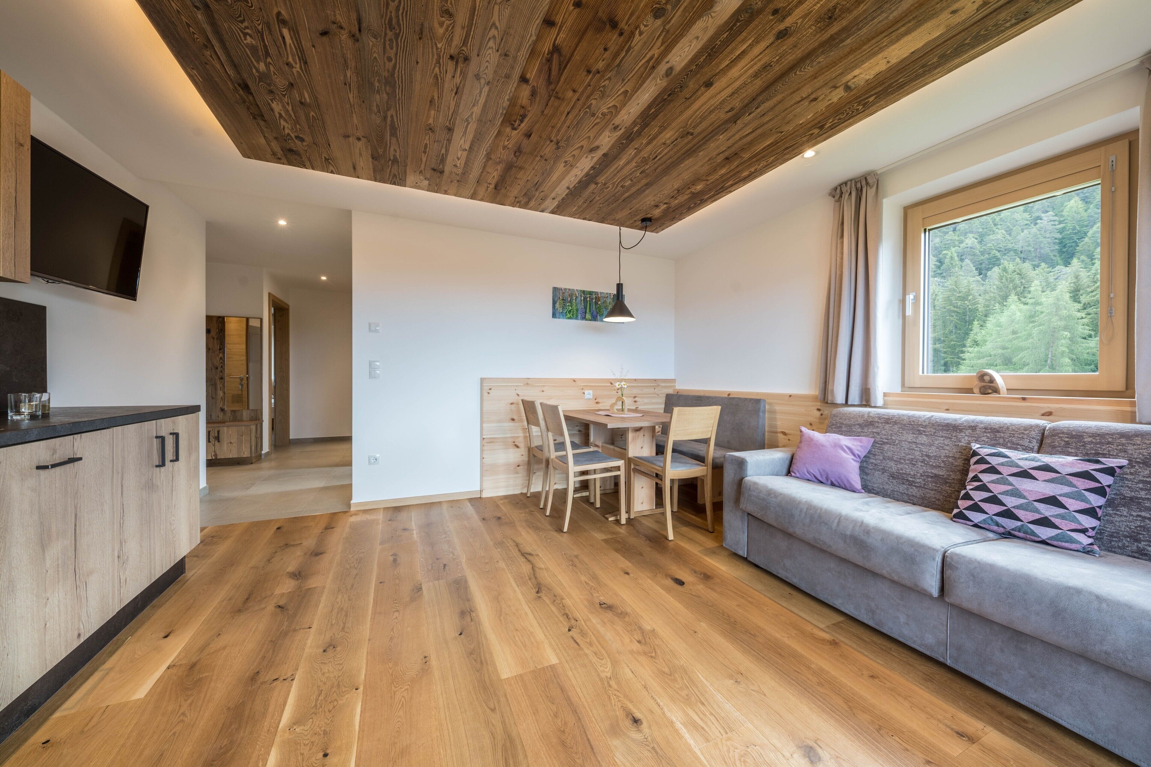 Et moderne interiør med træloft, spiseplads med bord og stole, sofa med puder, og et fjernsyn ophængt på væggen.