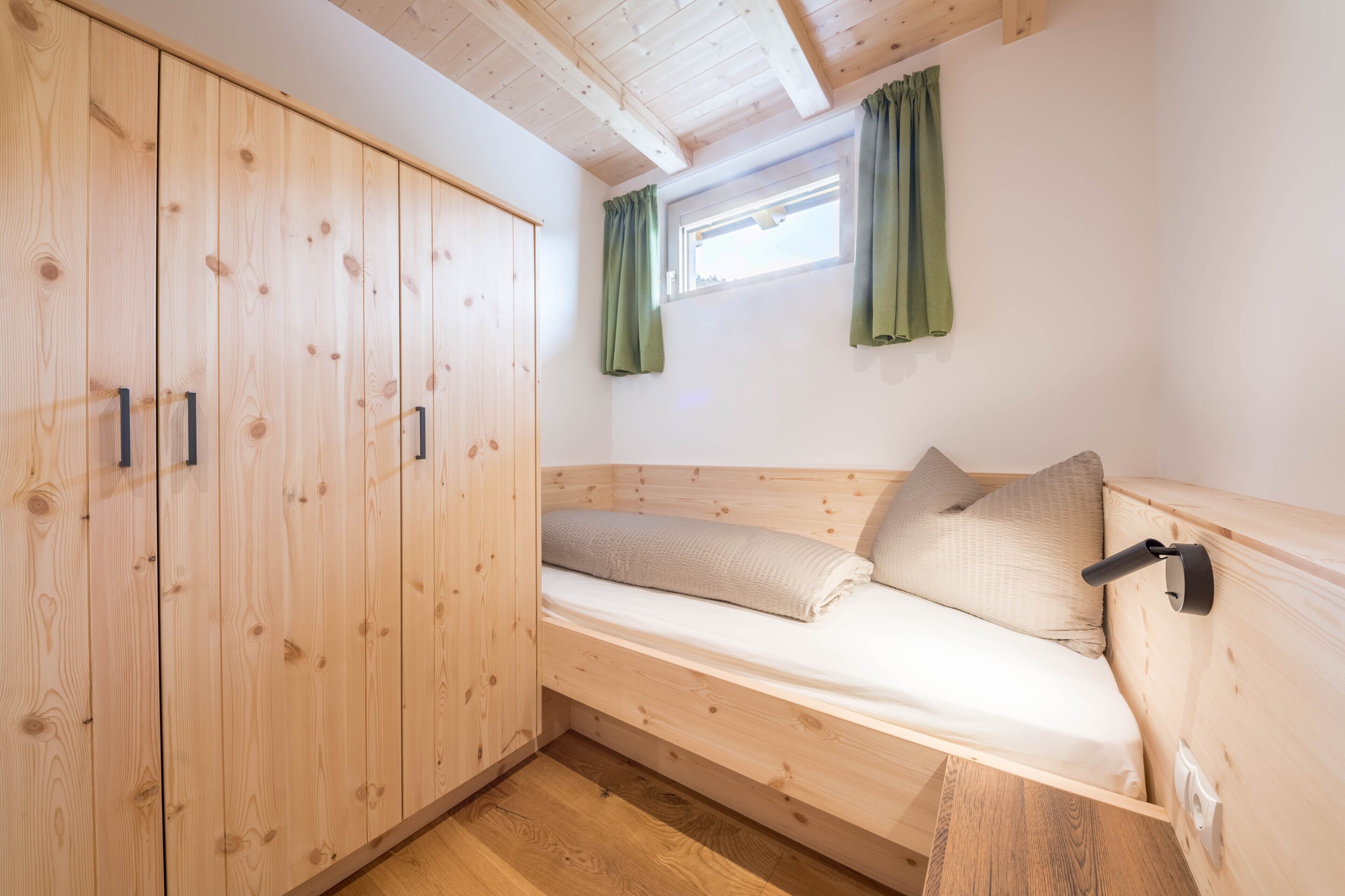 Uma cama de beliche de madeira com encosto, um guarda-roupa de madeira com duas portas e uma janela com cortinas.