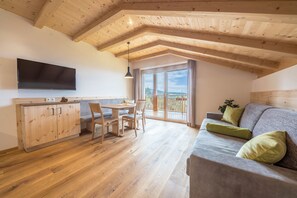 A room with a wooden ceiling, a flat-screen TV, a sofa with green cushions, a dining table with chairs, and a sliding glass door leading to a balcony.