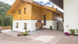 Casa de madera de dos pisos con un balcón, un coche estacionado frente a ella y montañas al fondo.