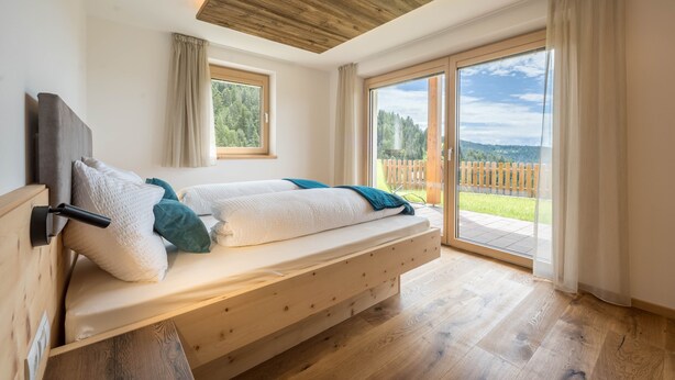 Ein Schlafzimmer mit einem Holzbett, einem Fenster und Blick auf eine Holzterrasse und Bäume.