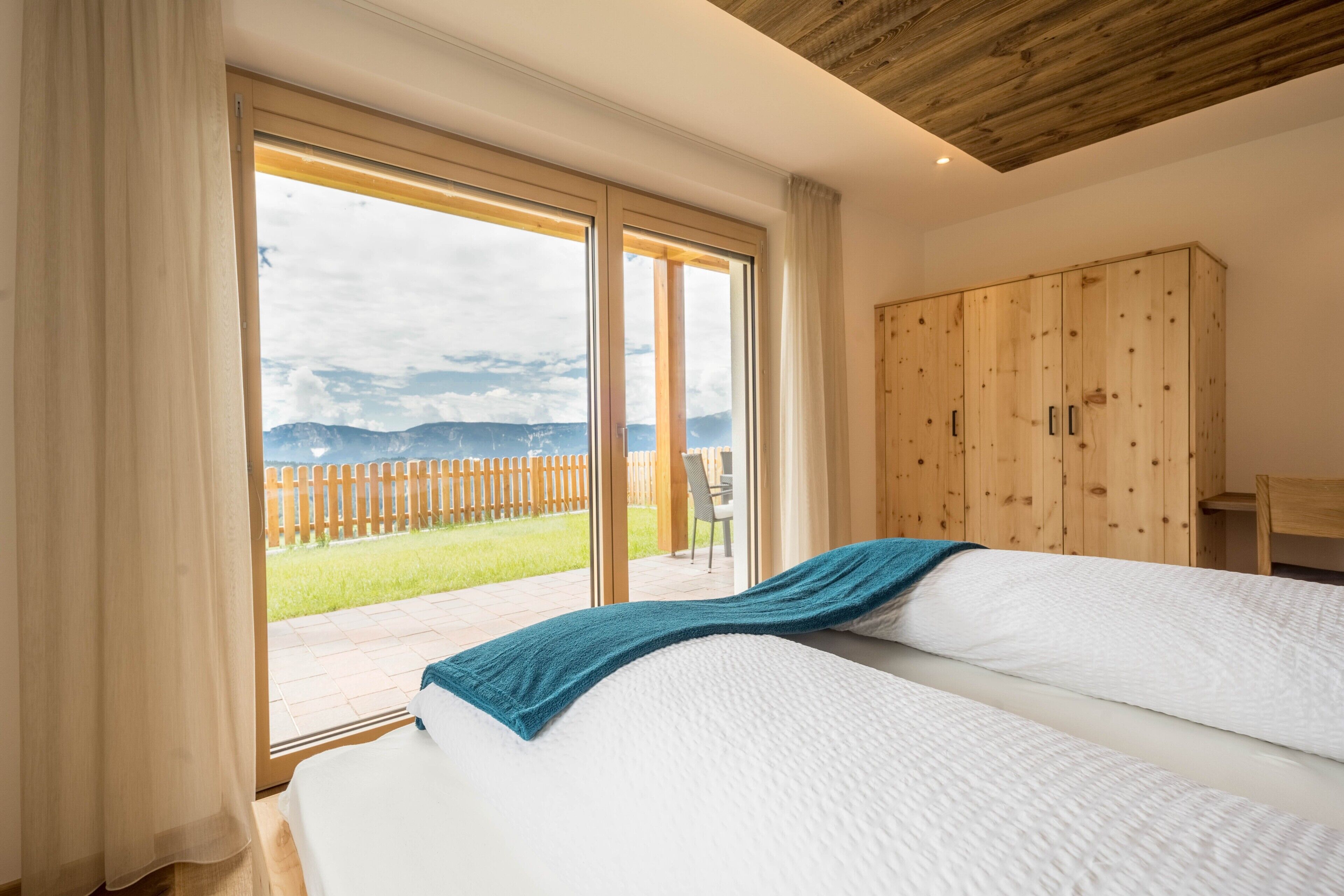 A bedroom with a large sliding glass door leading to a balcony, a wooden wardrobe, and a bed with white bedding.