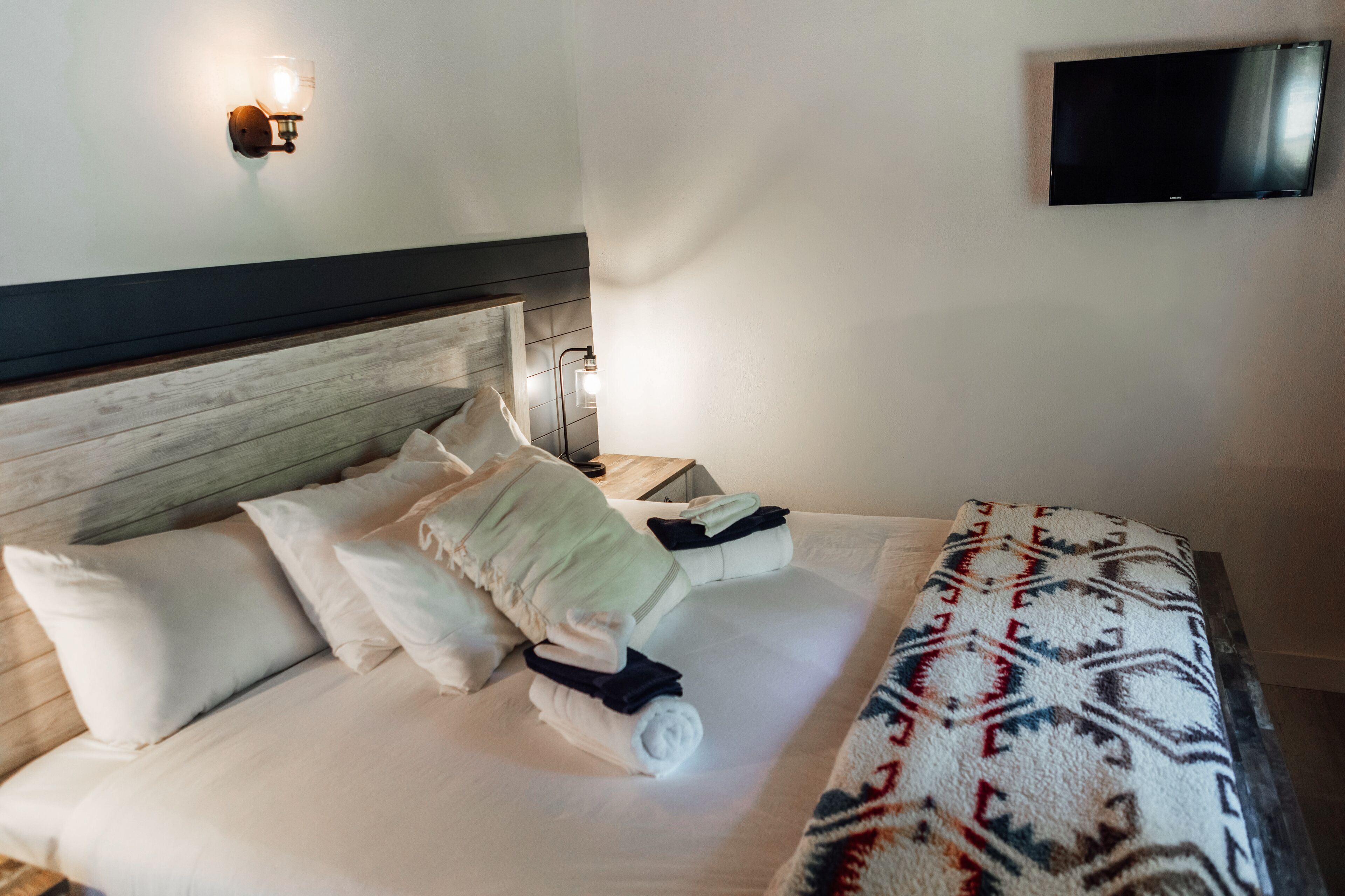 Un letto ben rifatto con cuscini e coperta, un comodino con una lampada e un televisore a schermo piatto montato a parete.