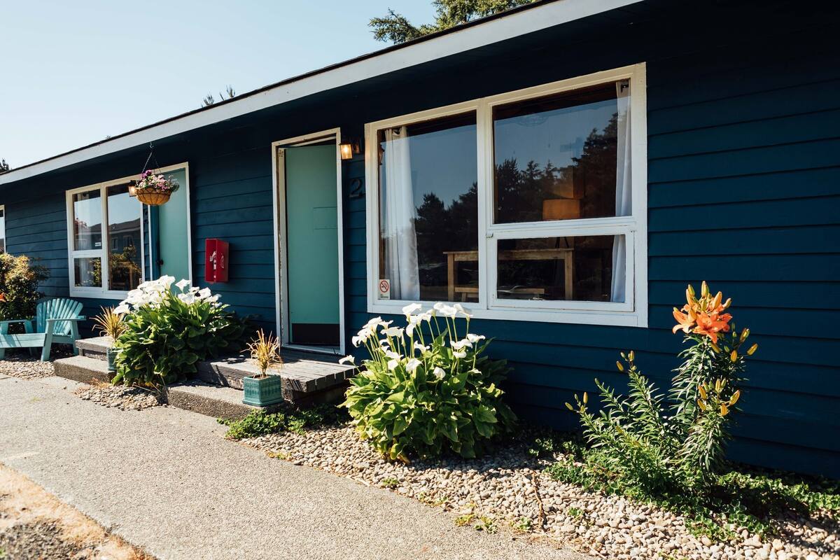 Ett blått hus med vitmålade fönster, en grön dörr och en liten veranda med en stol och krukväxter.