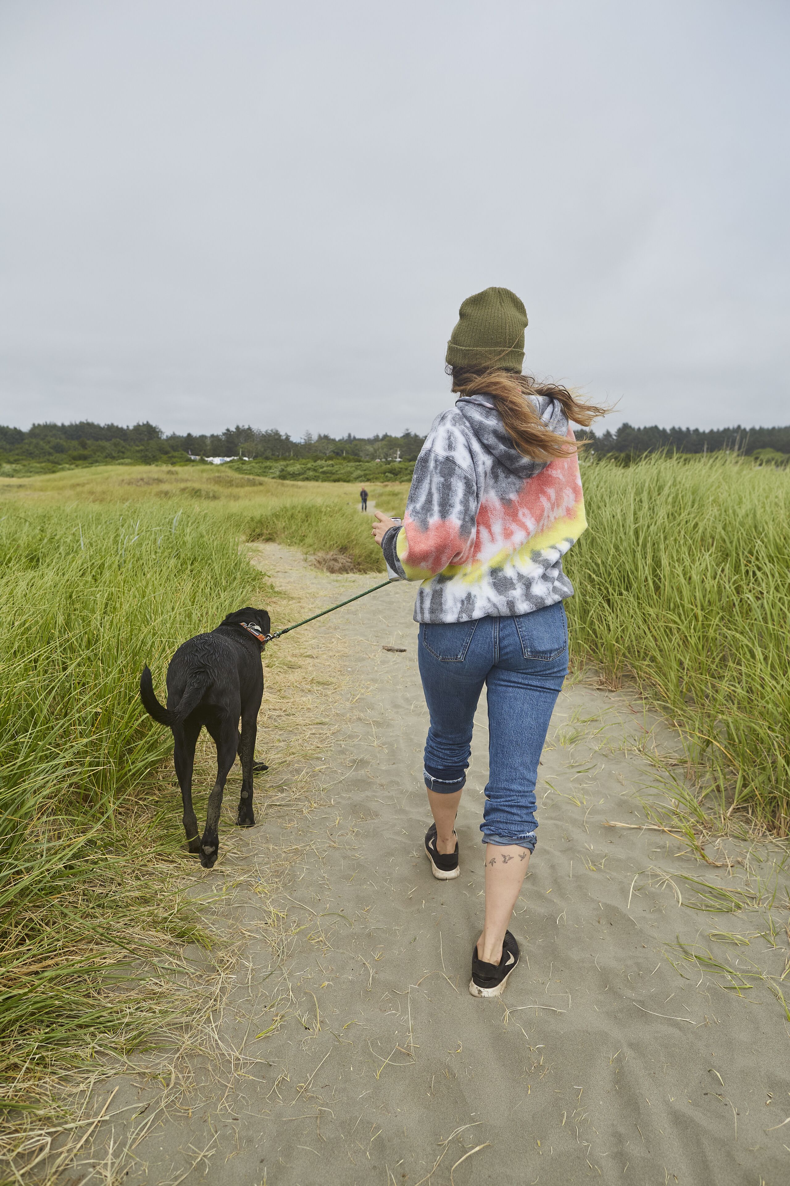 A person walking a dog on a sandy path with tall grass on either side.
