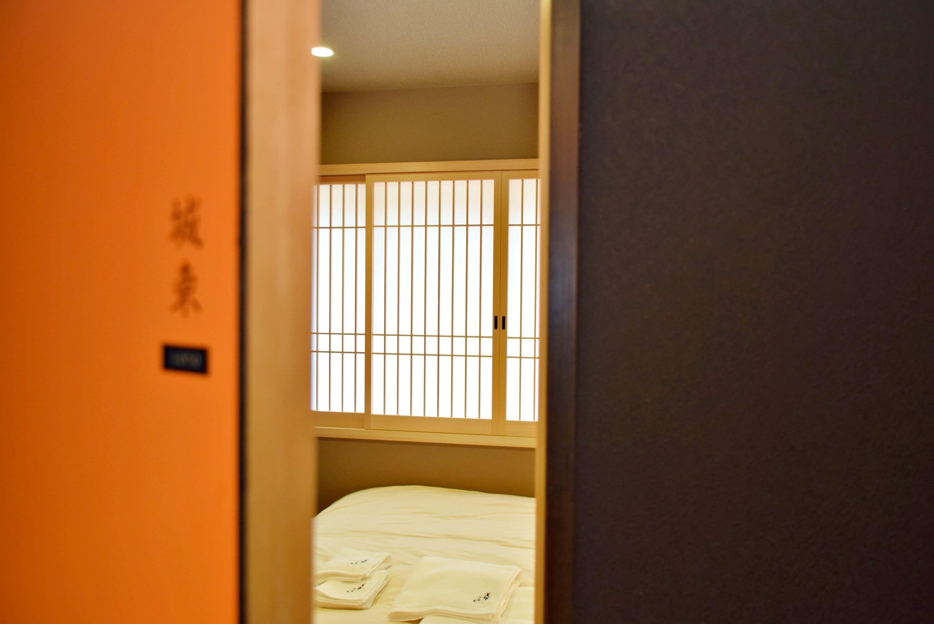Habitaci ón de hotel con una cama, una ventana con persianas verticales y una puerta con pared naranja y caracteres japoneses.