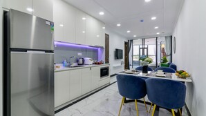 A modern kitchen with a stainless steel refrigerator, white cabinets, and a dining area with blue chairs and a table. - Aura Apartment Da Lat (Xuan Huong - Da Lat)