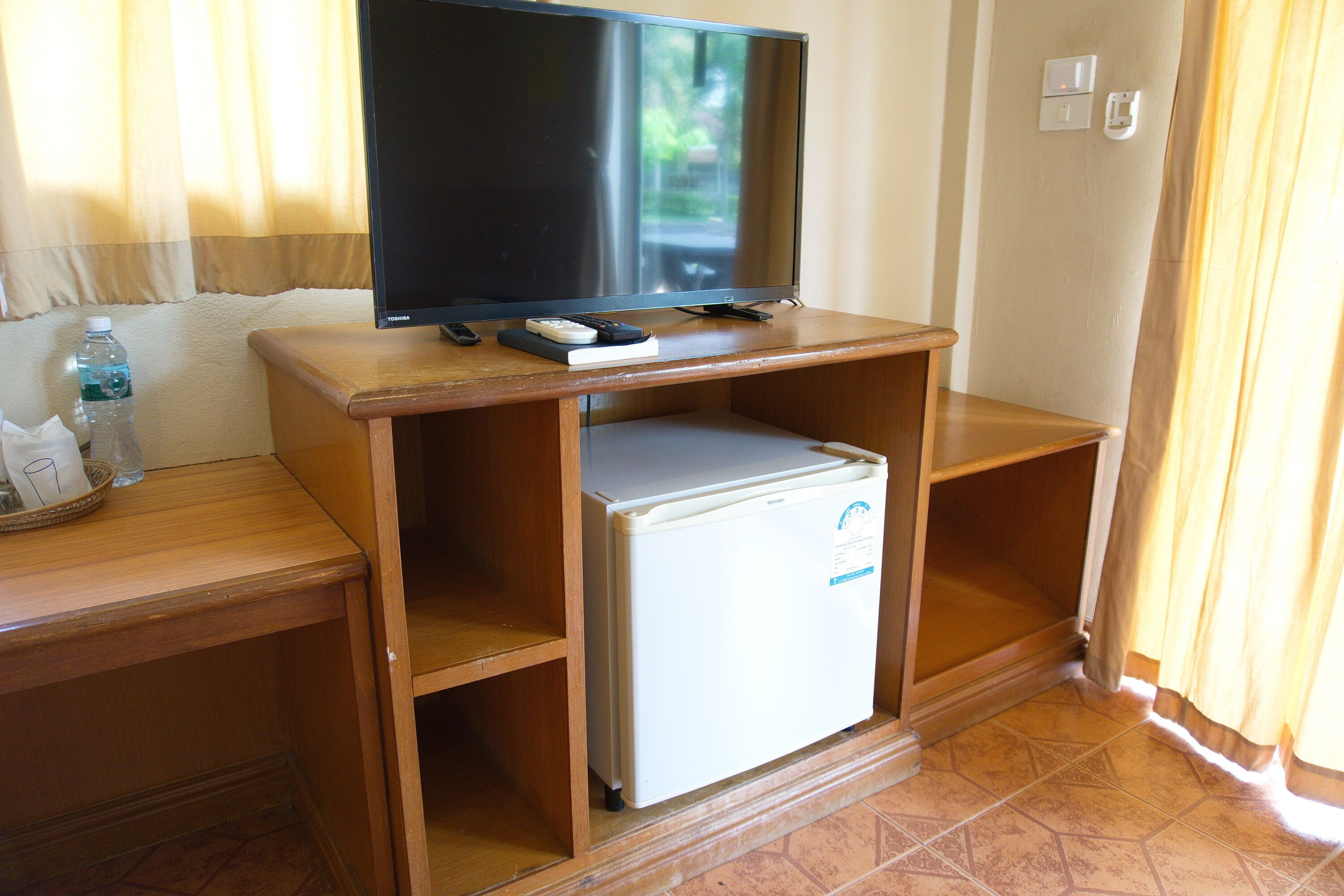 A compact room with a television on a wooden stand, a small refrigerator, and a wooden desk with a bottle of water and a tissue box.