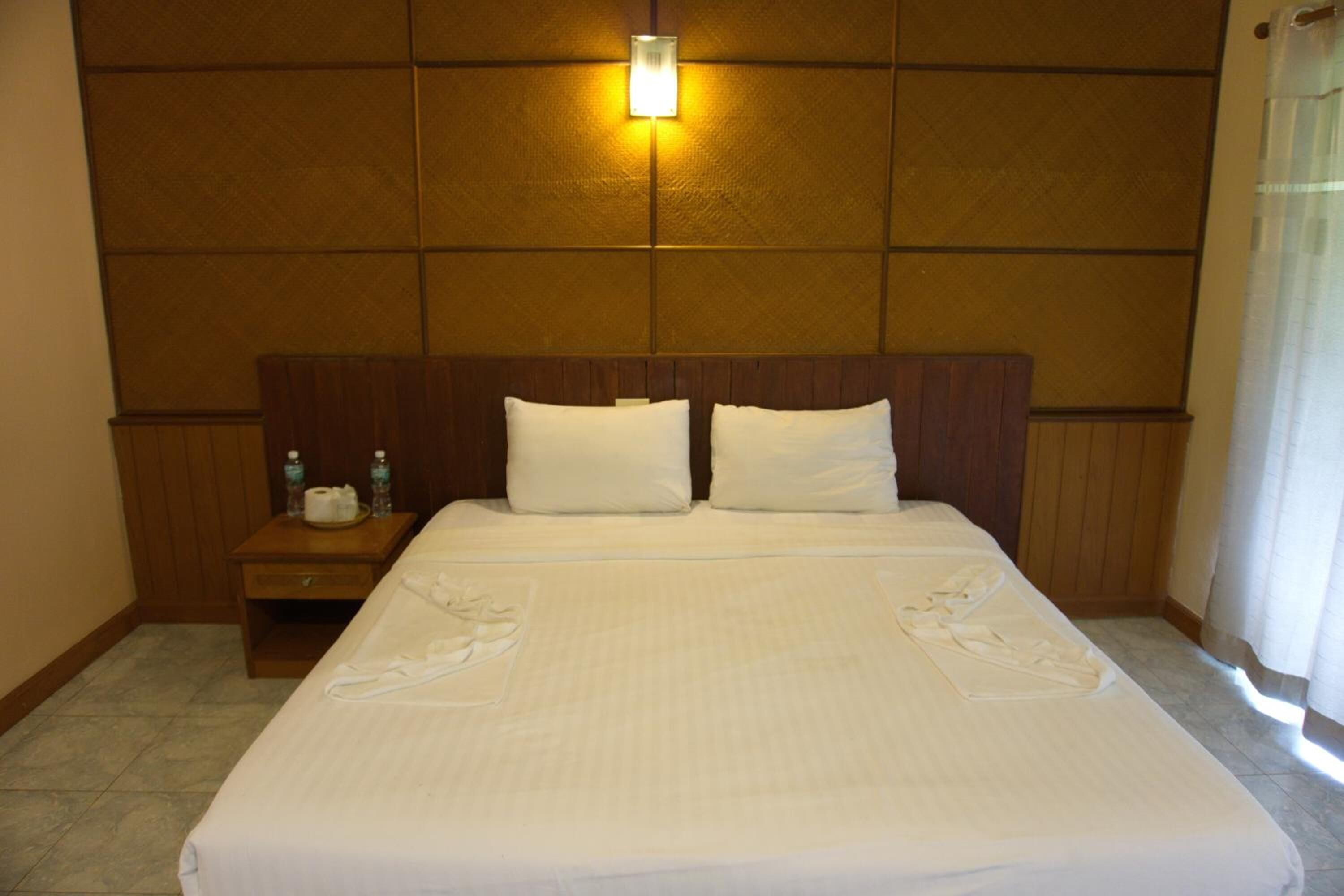 A hotel room with a large bed, two pillows, a nightstand with toiletries, and a wall-mounted light.